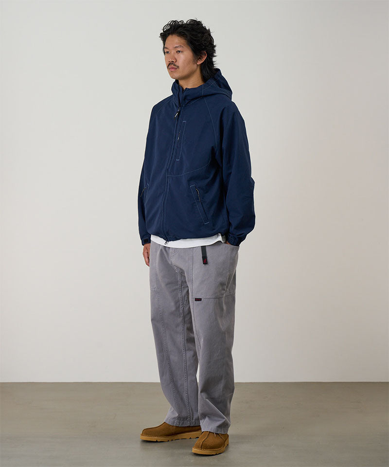 Model wearing a natural dyed grey Gramicci Gadget Pants and blue jacket on a light-tone background.
