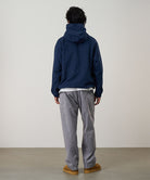 Back view of a model wearing a natural dyed grey Gramicci Gadget Pants and blue jacket on a light-tone background.
