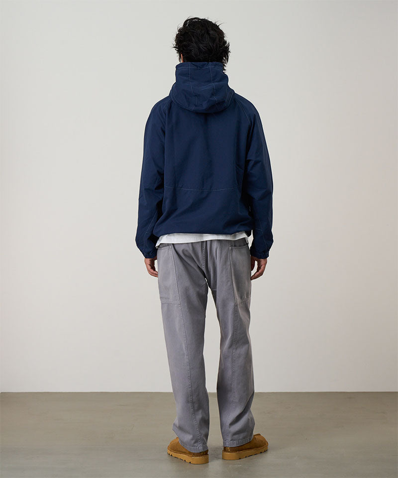 Back view of a model wearing a natural dyed grey Gramicci Gadget Pants and blue jacket on a light-tone background.

