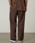 Model wearing walnut Gramicci Milton Pants and a brown shirt on a light-tone background.
