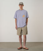 Man wearing a light purple t-shirt with a logo and beige shorts against a plain background