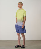Model wearing a yellow gradient Gramicci UPF Shield S/S Top with a white Gramicci graphic and blue shorts on a light-tone background.
