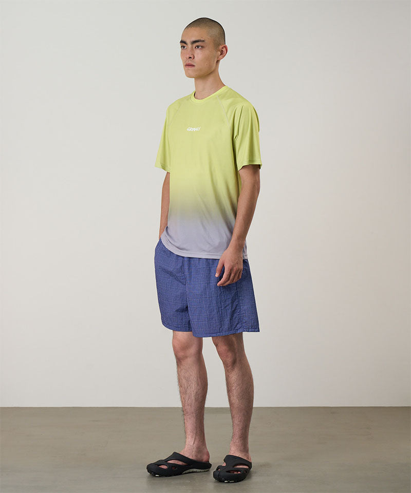 Model wearing a yellow gradient Gramicci UPF Shield S/S Top with a white Gramicci graphic and blue shorts on a light-tone background.
