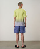 Back view of a model wearing a yellow gradient Gramicci UPF Shield S/S Top with a white Gramicci graphic and blue shorts on a light-tone background.
