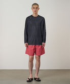 Model wearing a black Gramicci UPF Shield S/S Top with a sleeve graphic and red shorts on a light-tone background.
