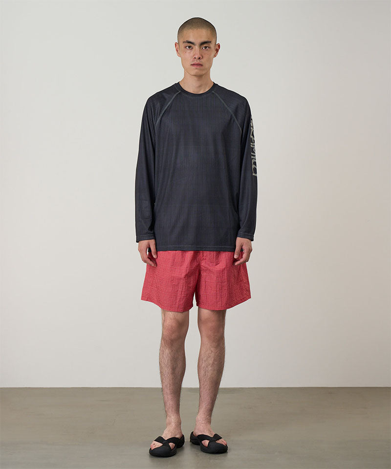 Model wearing a black Gramicci UPF Shield S/S Top with a sleeve graphic and red shorts on a light-tone background.
