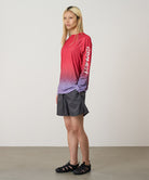 Model wearing a red gradient Gramicci UPF Shield S/S Top with a sleeve graphic and black shorts on a light-tone background.
