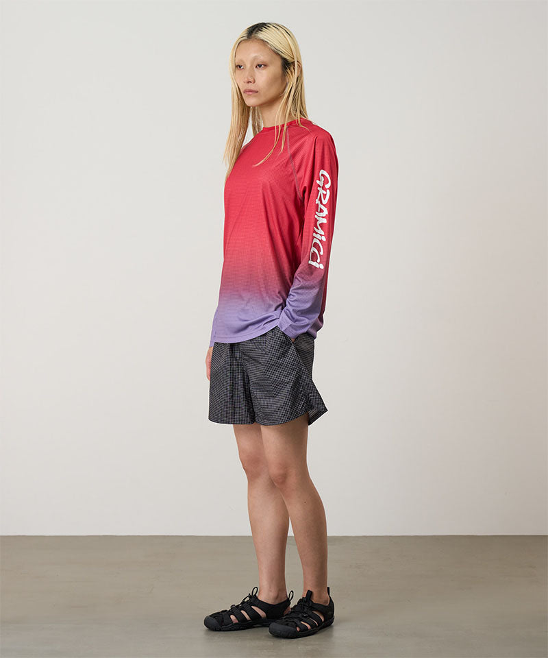 Model wearing a red gradient Gramicci UPF Shield S/S Top with a sleeve graphic and black shorts on a light-tone background.
