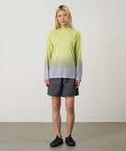 Model wearing a yellow gradient Gramicci UPF Shield S/S Top with a sleeve graphic and black shorts on a light-tone background.

