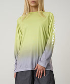 Model wearing a yellow gradient Gramicci UPF Shield S/S Top with a sleeve graphic and black shorts on a light-tone background.
