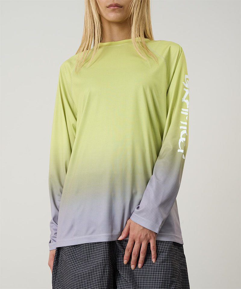 Model wearing a yellow gradient Gramicci UPF Shield S/S Top with a sleeve graphic and black shorts on a light-tone background.
