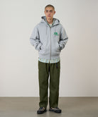 Person wearing a gray hoodie with a logo, plaid shirt, and green pants against a plain background