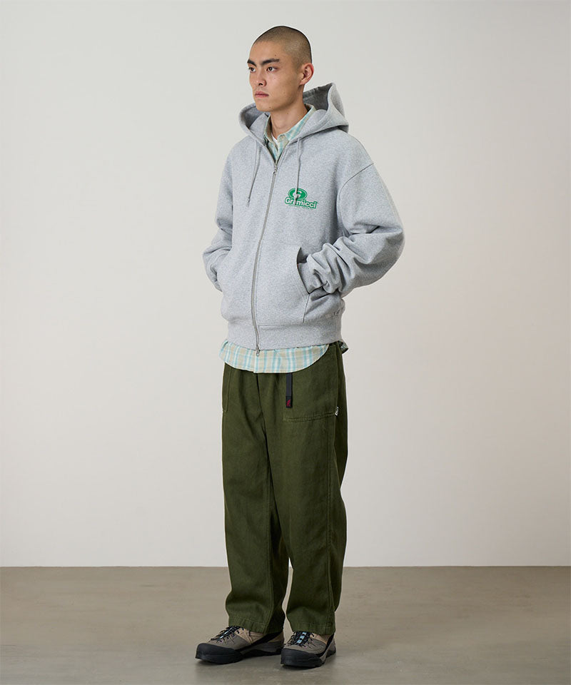 Person wearing a gray hoodie with a logo and green pants against a plain background