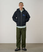 Person wearing a dark hoodie with a logo and green pants against a plain background