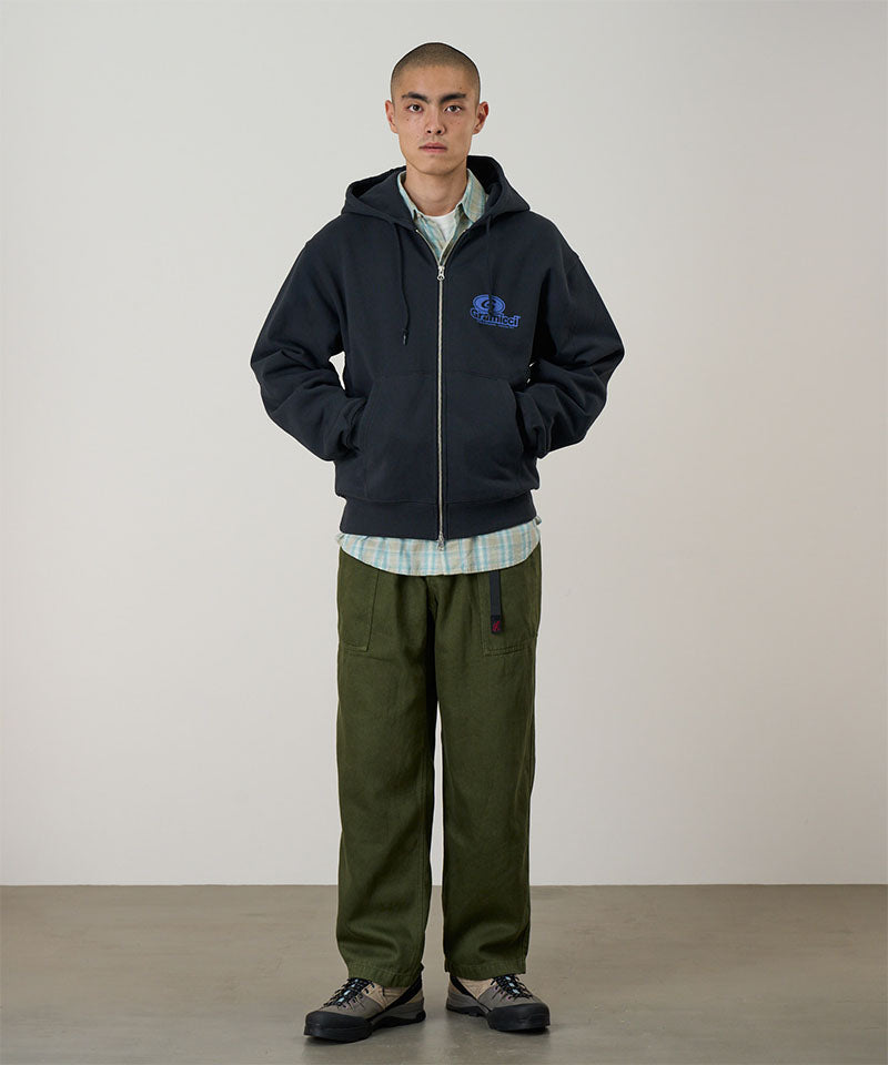 Person wearing a dark hoodie with a logo and green pants against a plain background