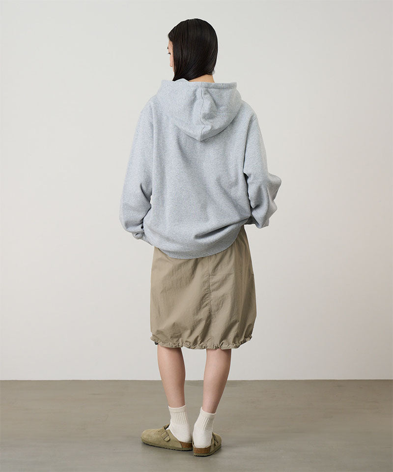 Person wearing a light gray hoodie and beige skirt standing against a plain background