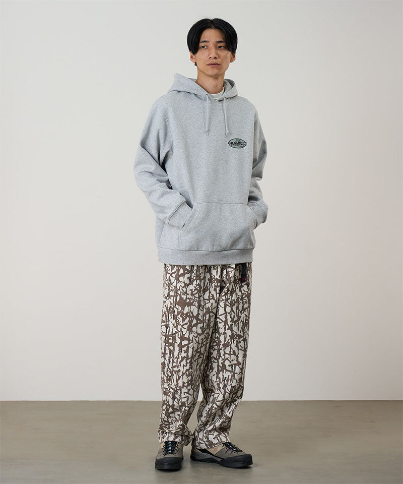 Person wearing a gray hoodie and patterned pants standing against a plain background