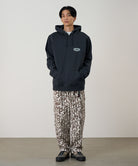 Person wearing a dark hoodie and patterned pants standing against a plain background