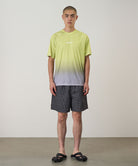 Model wearing black checked Gramicci Grid Swim Shorts and a lime and grey gradient t-shirt on a light-tone background.
