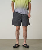 Model wearing black checked Gramicci Grid Swim Shorts and a lime and grey gradient t-shirt on a light-tone background.
