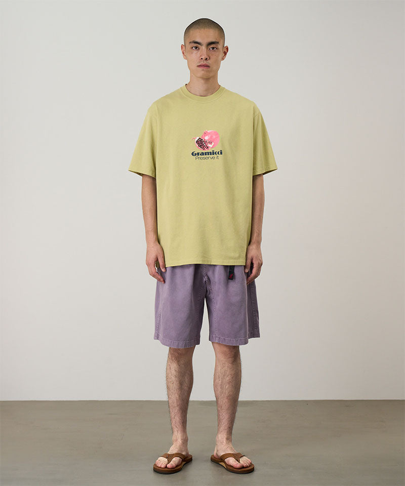 Model wearing a lime green Gramicci Pomegranate Tee with a a graphic and purple shorts on a light tone background.
