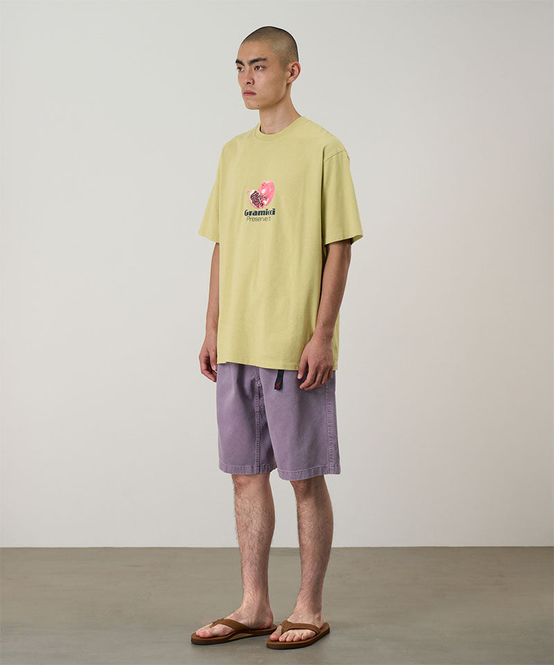 Model wearing a lime green Gramicci Pomegranate Tee with a a graphic and purple shorts on a light tone background.
