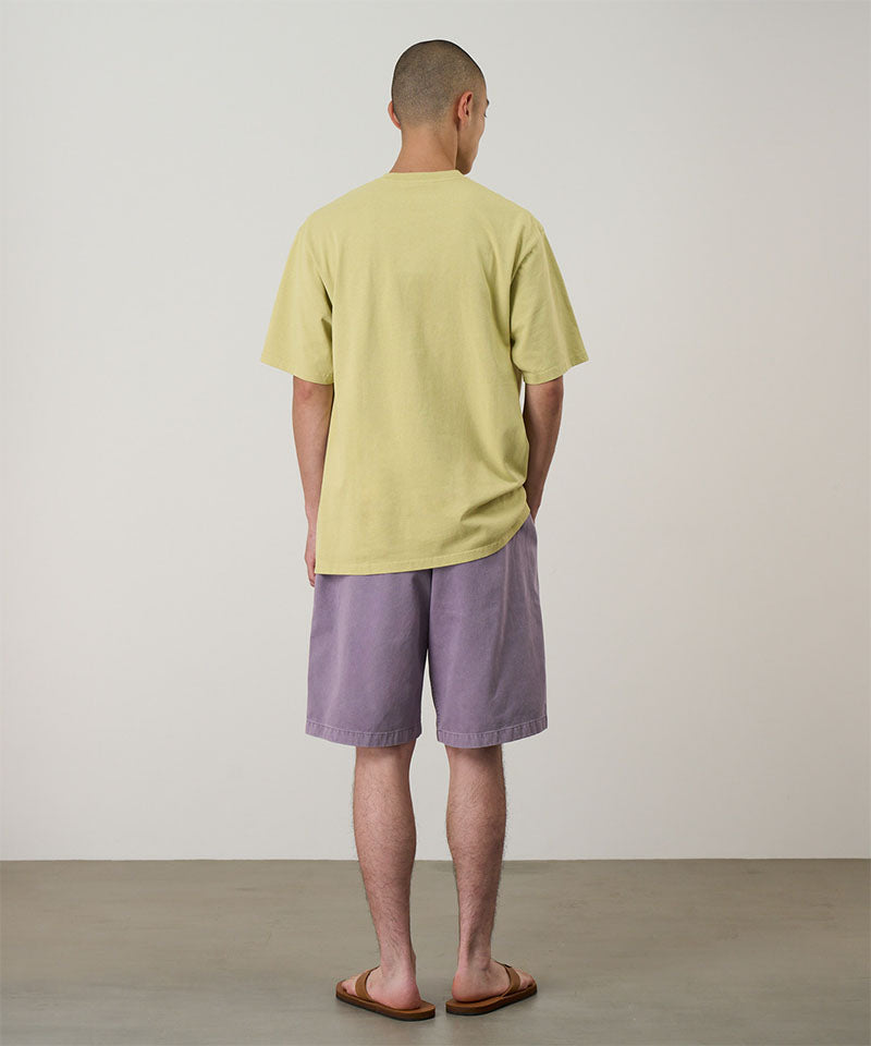 Back view of a model wearing a lime green Gramicci Pomegranate Tee and purple shorts on a light tone background.
