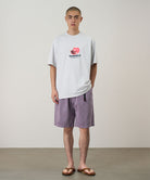 Model wearing a smoke colored Gramicci Pomegranate Tee with a a graphic and purple shorts on a light tone background.
