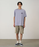 Model wearing a light lavender Gramicci '82 Tee with a dark lavender graphic and beige shorts on a light tone background.