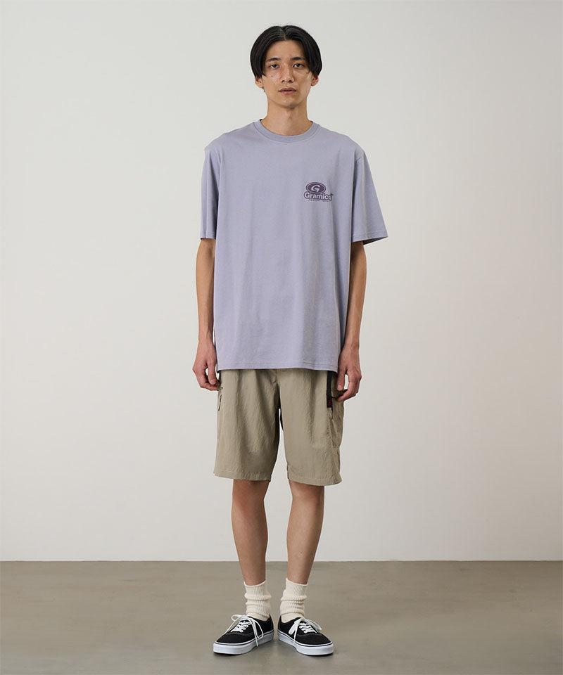 Model wearing a light lavender Gramicci '82 Tee with a dark lavender graphic and beige shorts on a light tone background.