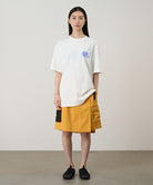 Model wearing a white Gramicci '82 Tee with a blue graphic and yellow skort on a light tone background.