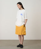 Model wearing a white Gramicci '82 Tee with a blue graphic and yellow skort on a light tone background.