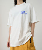 Model wearing a Gramicci white '82 Tee with blue graphic against a light tone background.