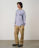 Model wearing a lavender Gramicci Equipped For Dirt L/S Tee with a a Gramicci gear graphic graphic and olive shorts on a light tone background.