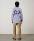 Rear view of a model wearing a lavender Gramicci Equipped For Dirt L/S Tee with a a Gramicci gear graphic graphic and olive shorts on a light tone background.