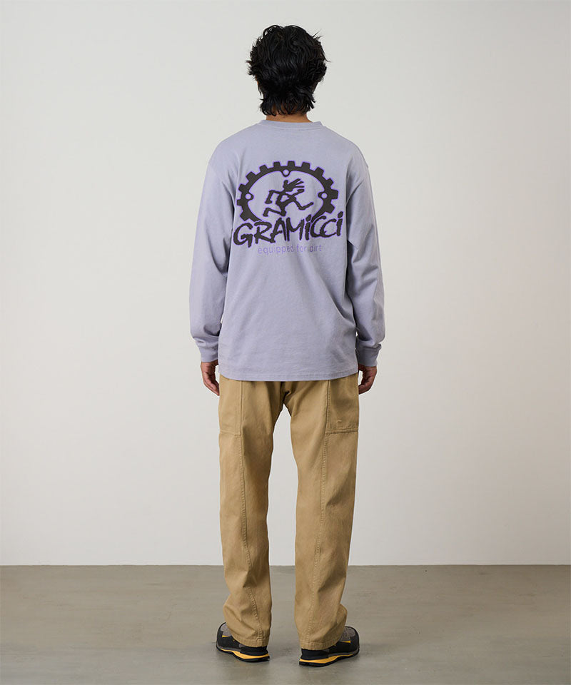 Rear view of a model wearing a lavender Gramicci Equipped For Dirt L/S Tee with a a Gramicci gear graphic graphic and olive shorts on a light tone background.