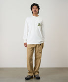Model wearing a white Gramicci Equipped For Dirt L/S Tee with a green graphic and chino colored pants on a light tone background.