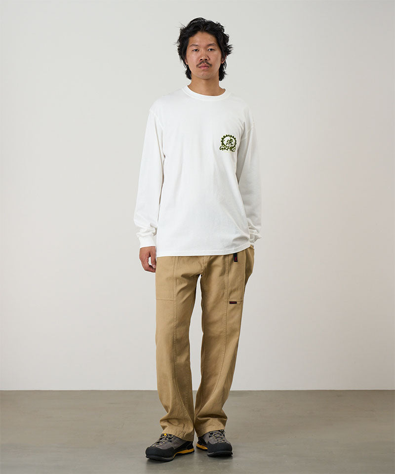 Model wearing a white Gramicci Equipped For Dirt L/S Tee with a green graphic and chino colored pants on a light tone background.