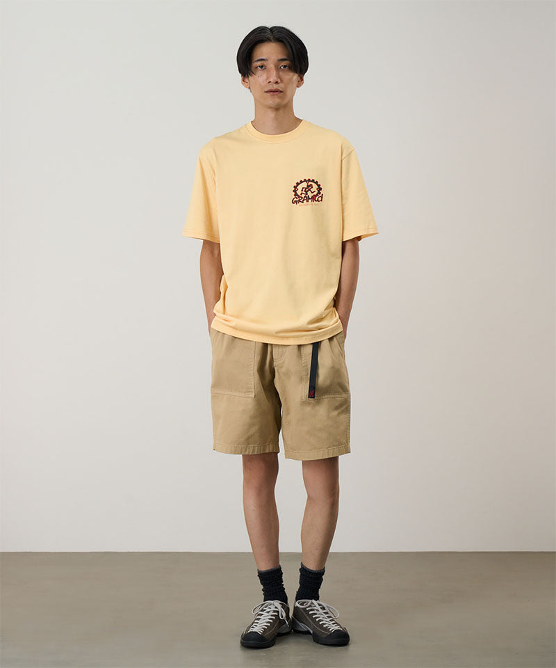 Model wearing a mango colored Gramicci Equipped For Dirt Tee with a graphic and beige shorts on a light-tone background.