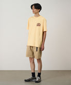 Model wearing a mango colored Gramicci Equipped For Dirt Tee with a graphic and beige shorts on a light-tone background.