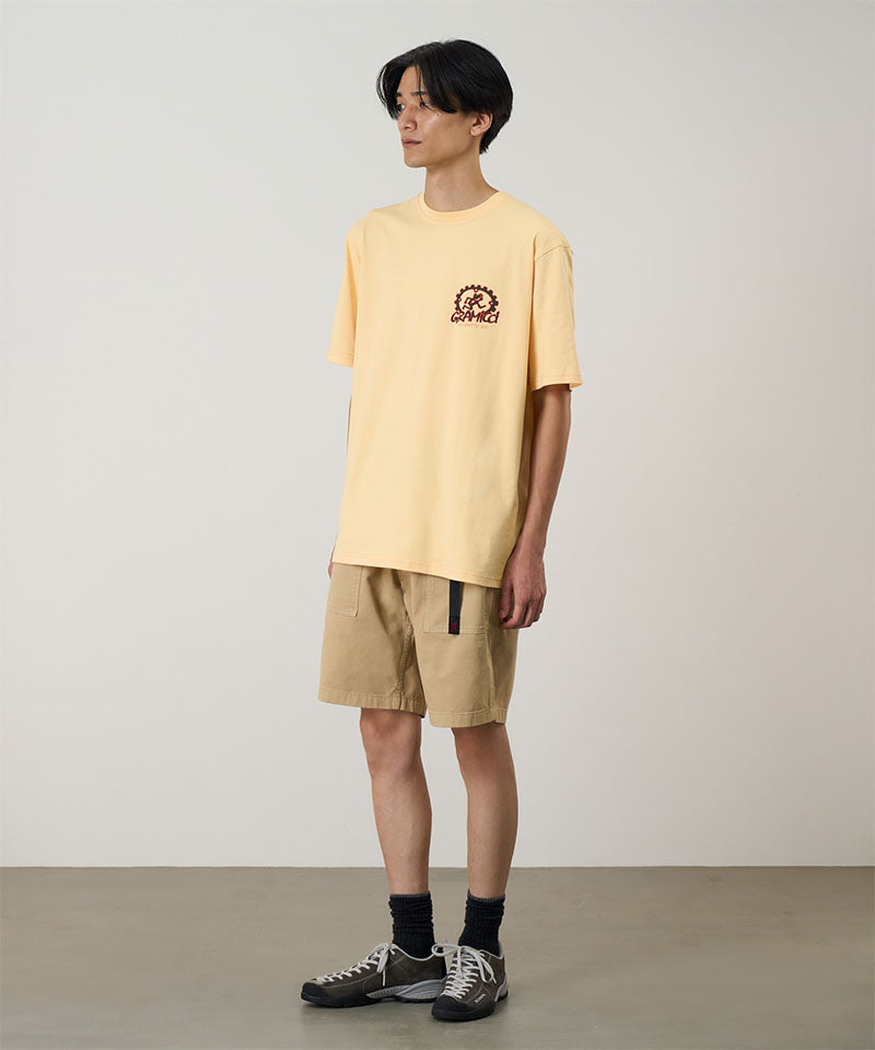 Model wearing a mango colored Gramicci Equipped For Dirt Tee with a graphic and beige shorts on a light-tone background.