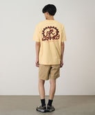Back view of a model wearing a mango colored Gramicci Equipped For Dirt Tee with a graphic and beige shorts on a light-tone background.