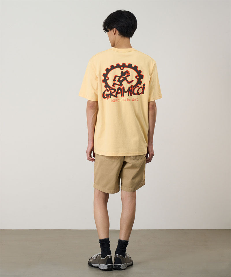 Back view of a model wearing a mango colored Gramicci Equipped For Dirt Tee with a graphic and beige shorts on a light-tone background.