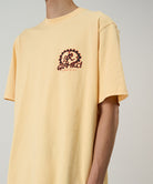 Model wearing a Gramicci mango colored Equipped For Dirt Tee with a graphic against a light-tone background.