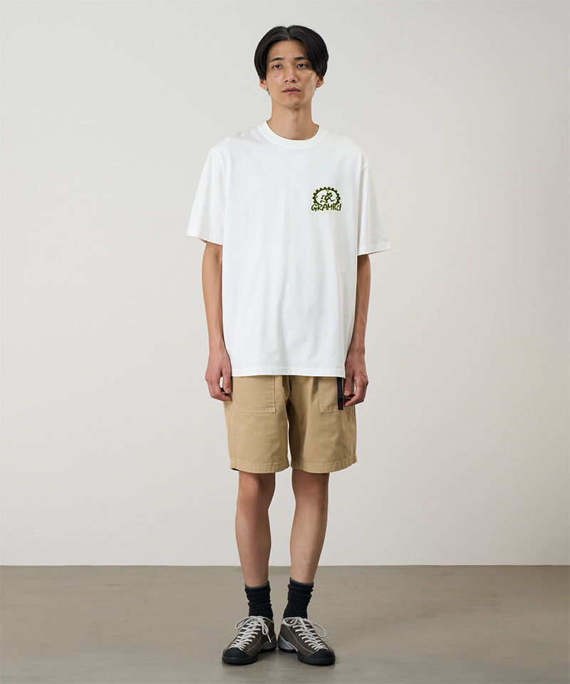 Model wearing a white Gramicci Equipped For Dirt Tee with a green graphic and beige shorts on a light-tone background.