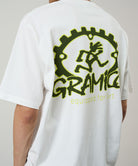 Back view of a model wearing a Gramicci white Equipped For Dirt Tee with a green graphic against a light-tone background.