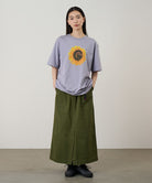 Model wearing a lavender Gramicci G-Flower Tee with a sunflower graphic and olive skirt on a light tone background.