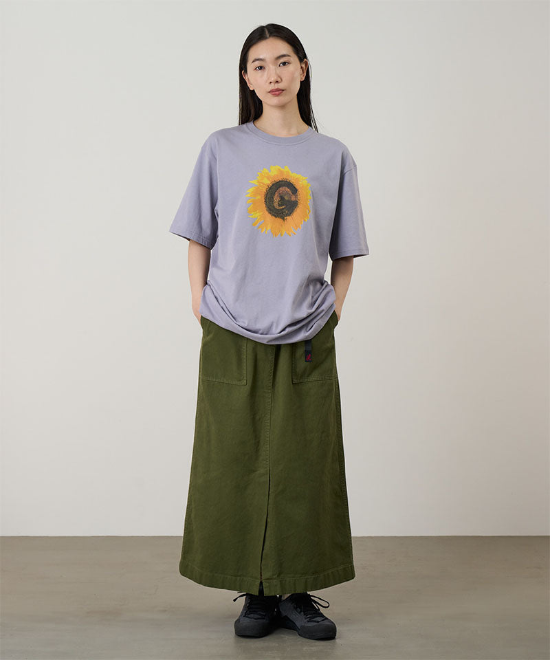 Model wearing a lavender Gramicci G-Flower Tee with a sunflower graphic and olive skirt on a light tone background.