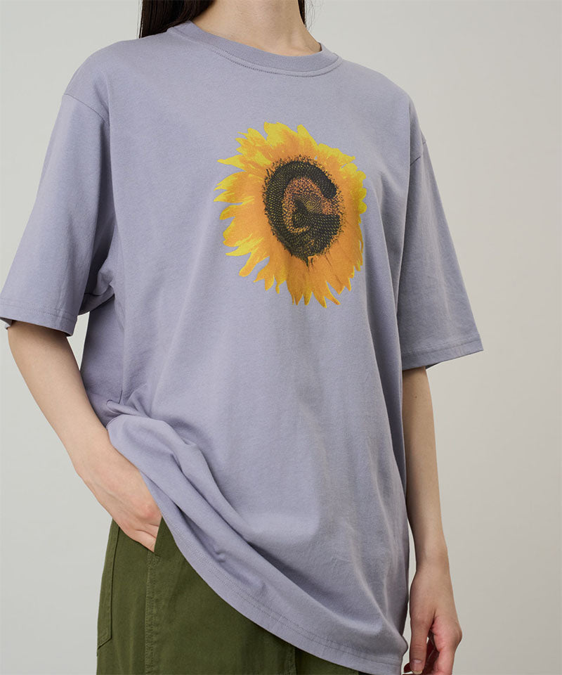 Model wearing a Gramicci lavender G-Flower Tee with sunflower graphic against a light tone background.
