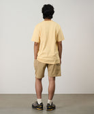 Back view of a model wearing a mango Gramicci G-Flower Tee with a sunflower graphic and beige shorts on a light tone background.